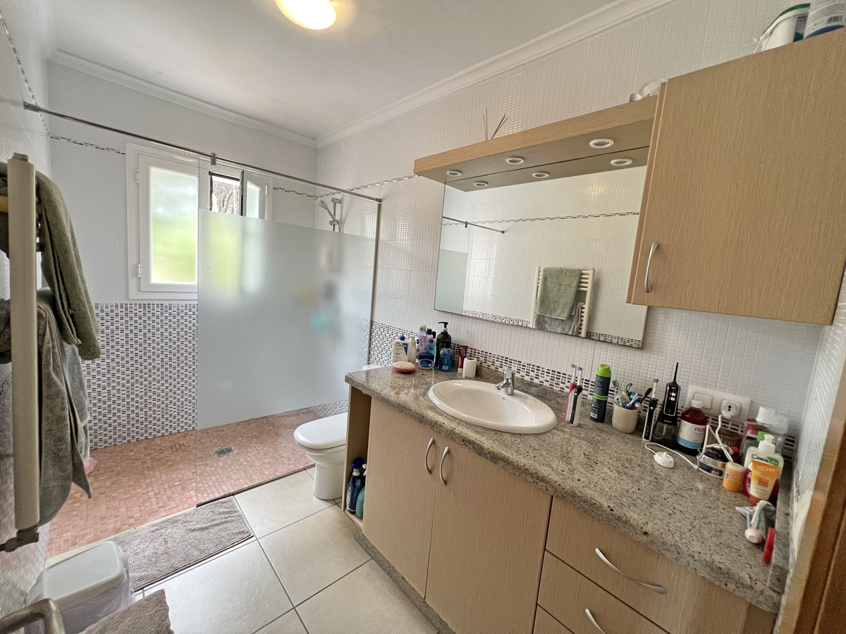 Salle de bain avec plan de travail en granit, meubles en bois clair et miroir à spots intégrés. Elle comprend une douche à l'italienne avec paroi en verre dépoli.
