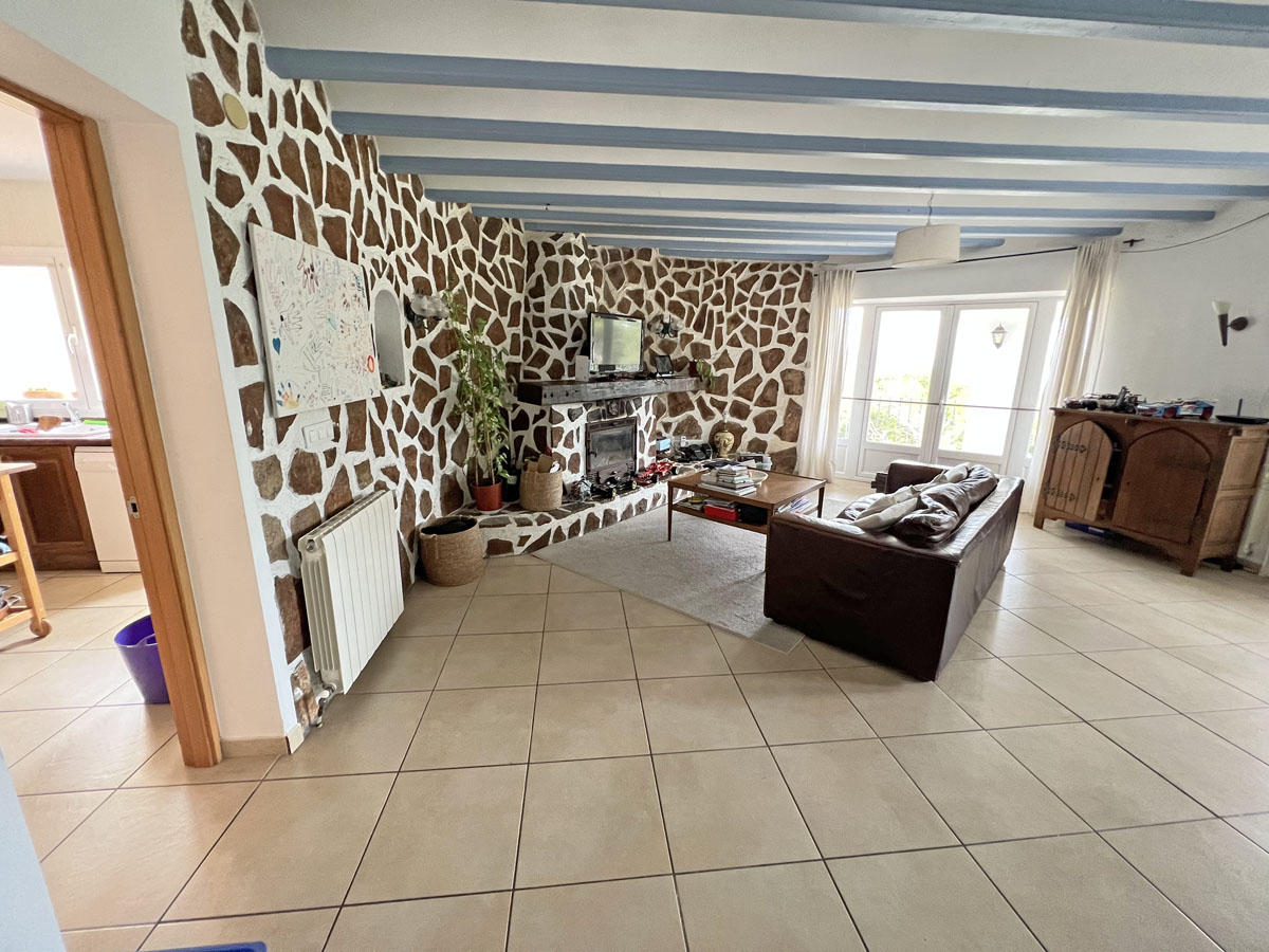 Séjour avec mur en pierre naturelle et cheminée, poutres apparentes teintées de bleu et accès direct à la terrasse par des portes vitrées.