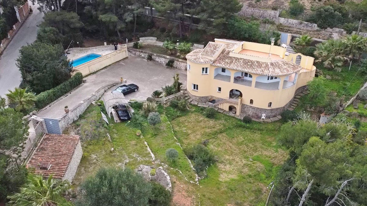Vue aérienne de la villa à façade courbe et soubassement en pierre, incluant une grande zone de stationnement, une piscine privée et un jardin méditerranéen arboré.