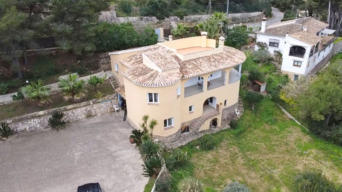 Vue aérienne de la villa méditerranéenne avec façade incurvée, toit en tuiles romaines et une grande zone de stationnement pavée entourée de jardins.