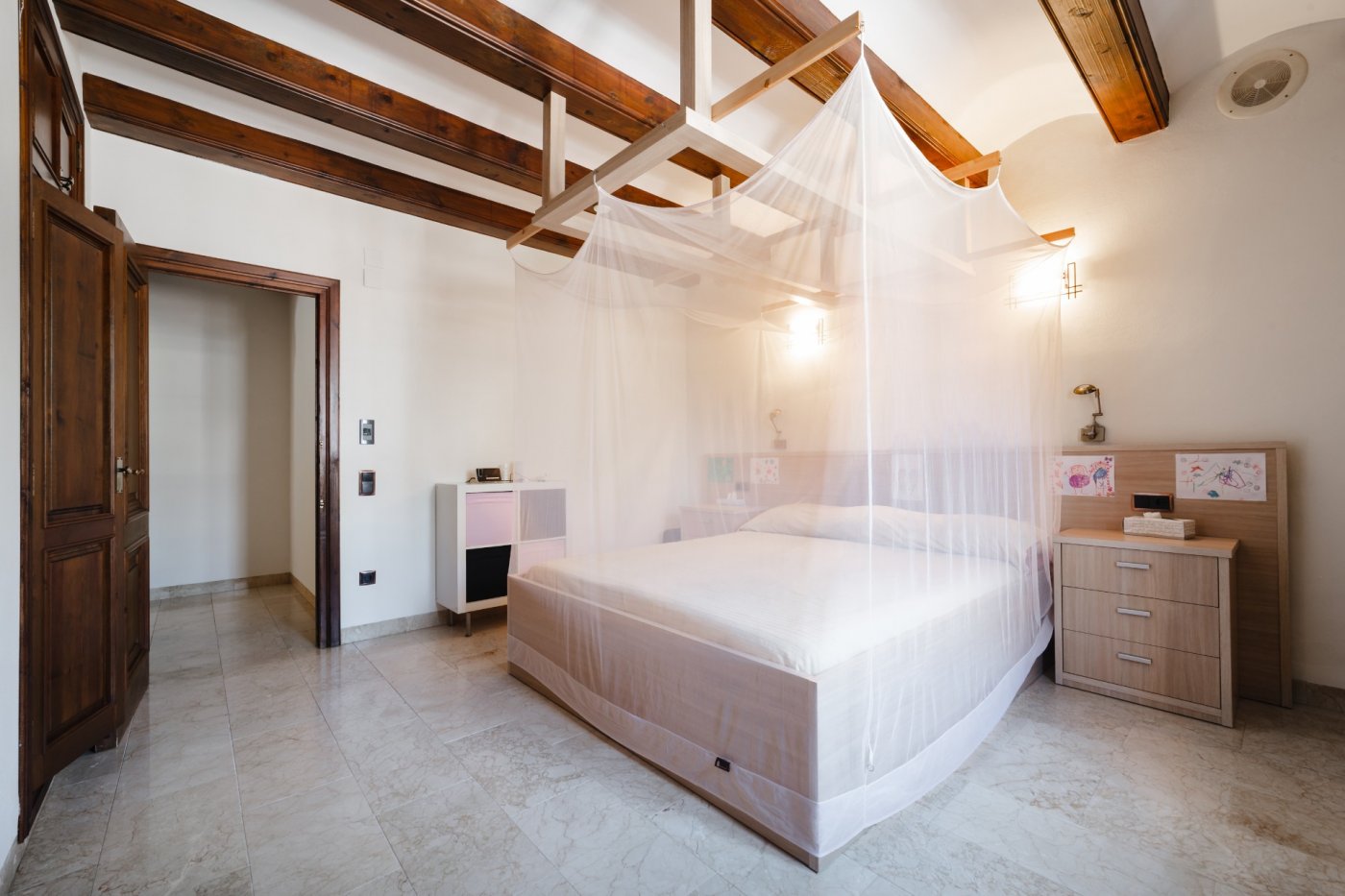 Chambre avec poutres apparentes en bois sombre, lit avec moustiquaire et sol en pierre naturelle. Portes en bois massif et éclairage mural fonctionnel.