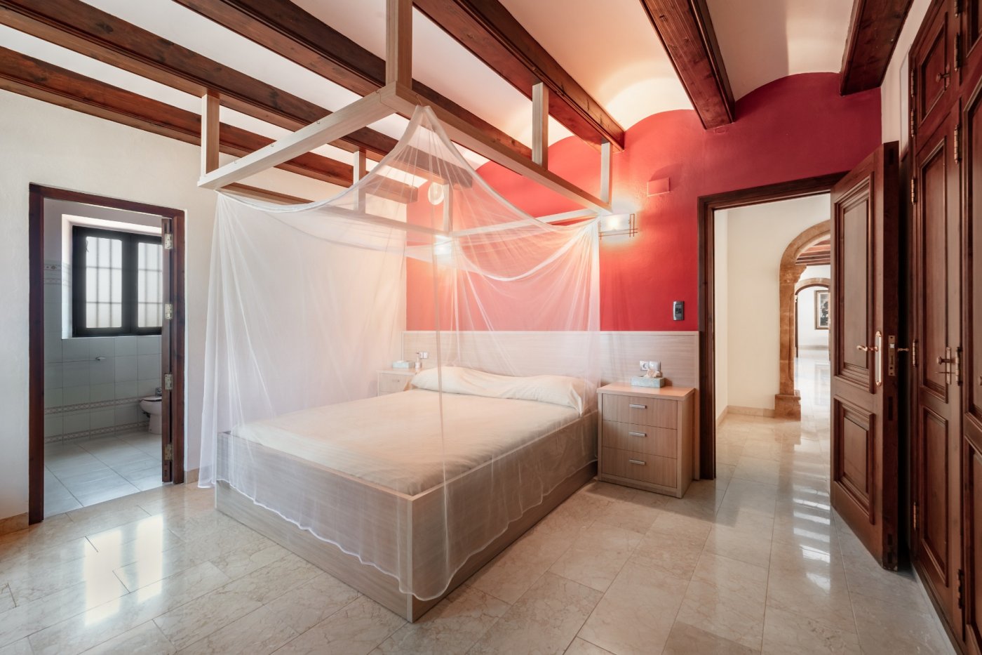 Chambre avec poutres apparentes, mur d'accent rouge et lit à baldaquin. Comprend un sol en marbre, une salle de bain attenante et un accès aux arches en pierre.