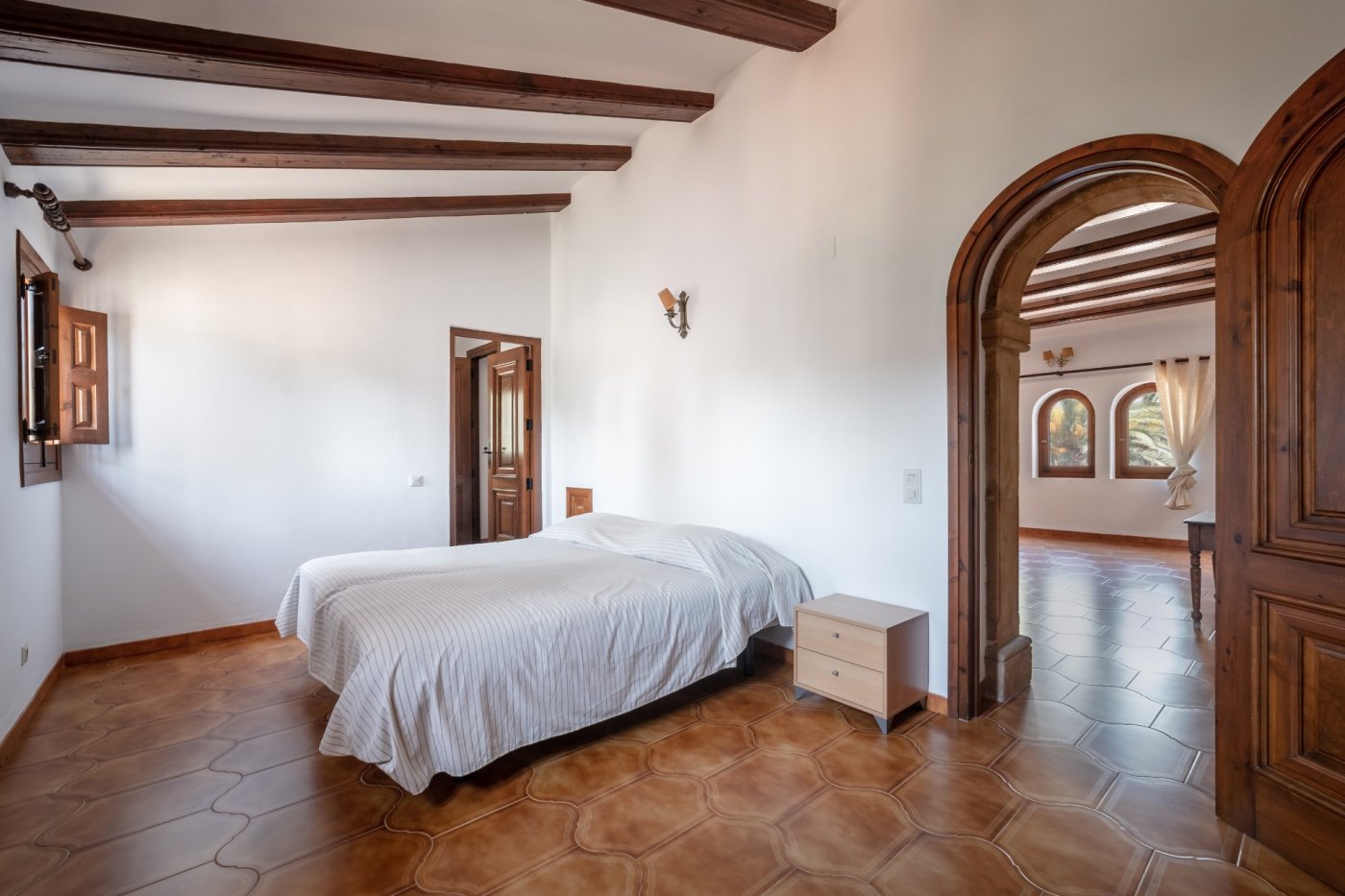 Chambre avec poutres en bois apparentes, sol en terre cuite et une arche en pierre de Tosca s'ouvrant sur une pièce lumineuse.