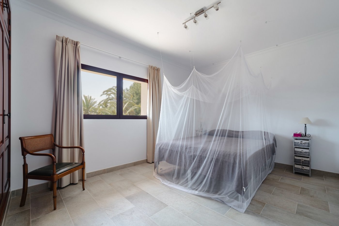 Chambre lumineuse avec lit double et moustiquaire suspendue, sol en grès cérame et fenêtre offrant une vue sur les palmiers du jardin méditerranéen.