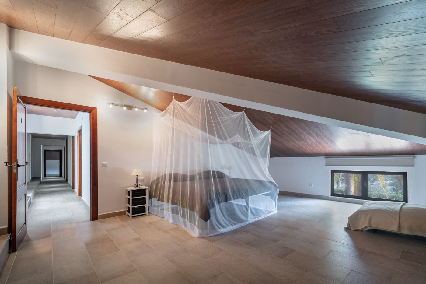 Chambre mansardée avec plafonds en bois, sol en grès cérame et lit avec moustiquaire. Une fenêtre basse donne sur le jardin et un couloir dessert l'étage.