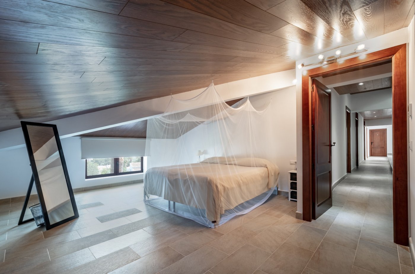 Chambre mansardée avec plafonds revêtus de bois, lit avec moustiquaire et carrelage au sol. La pièce comprend un miroir sur pied et s'ouvre sur un long couloir.
