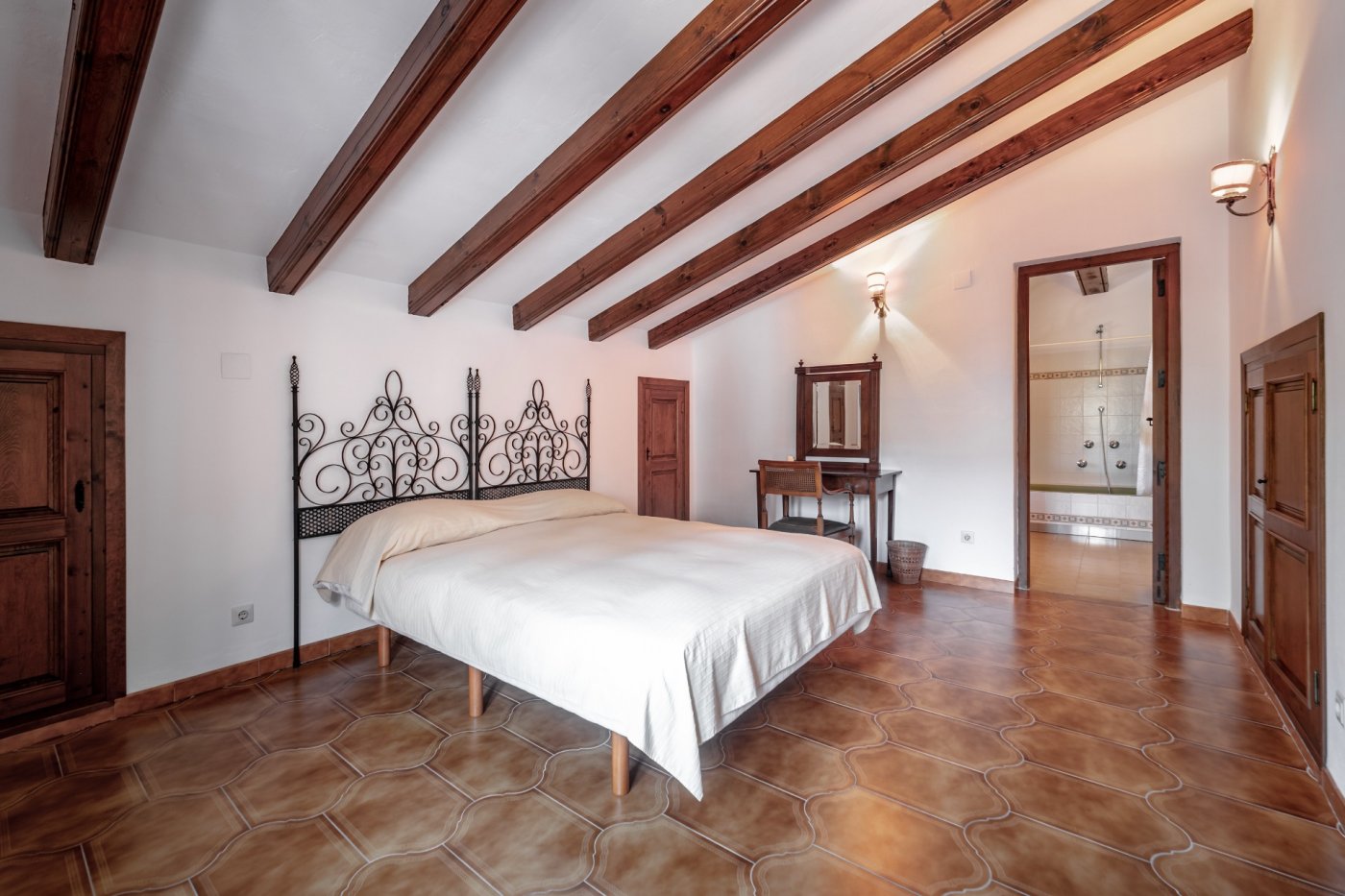 Chambre rustique avec poutres apparentes en bois, sol en terre cuite décorative, tête de lit en fer forgé et salle de bain attenante.