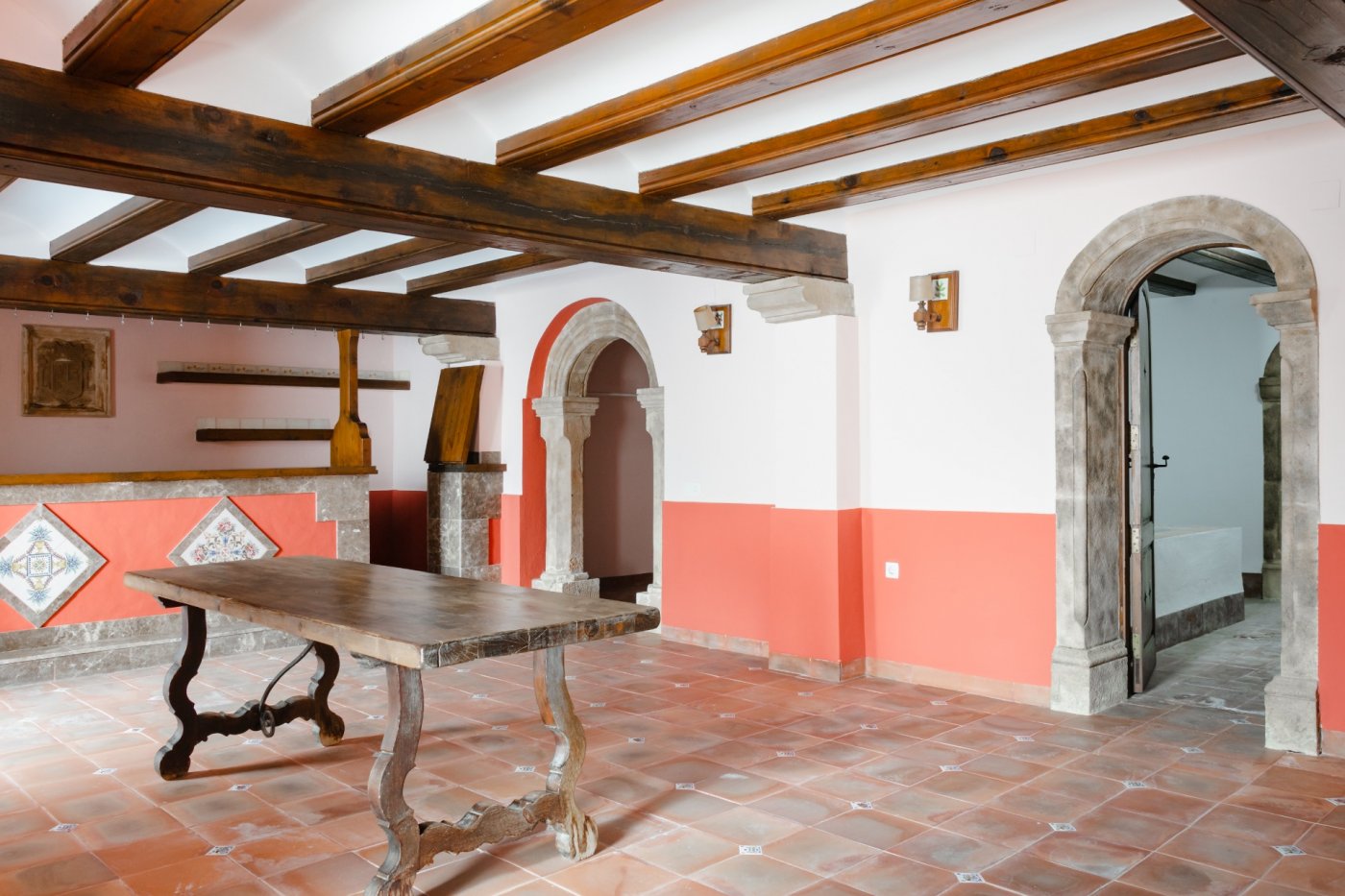 Espace de vie rustique avec poutres en bois apparentes, sol en terre cuite et arches en pierre de Tosca. Une table en bois massif souligne le style méditerranéen traditionnel.