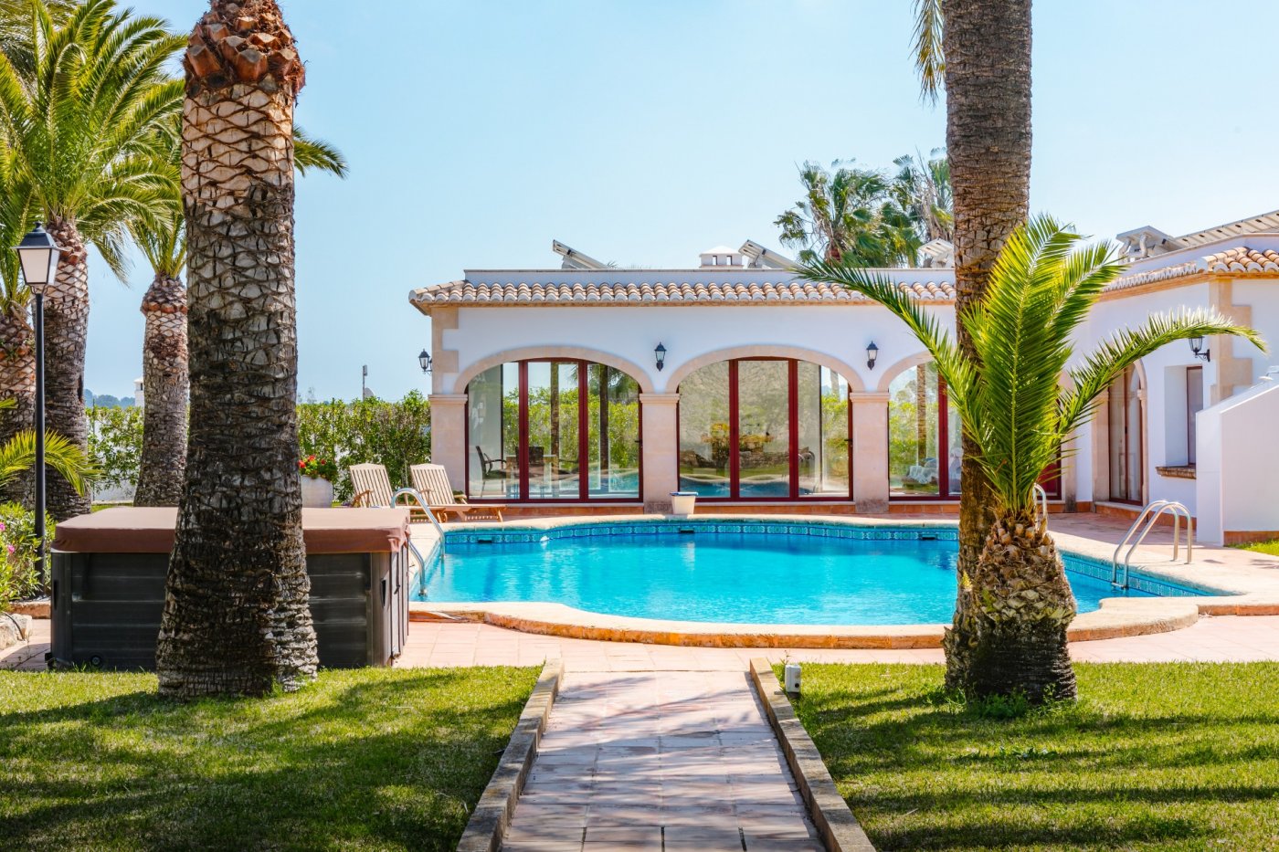 Espace piscine entouré de palmiers et de pelouse, avec une terrasse en terre cuite et une villa aux arches en pierre et panneaux photovoltaïques.