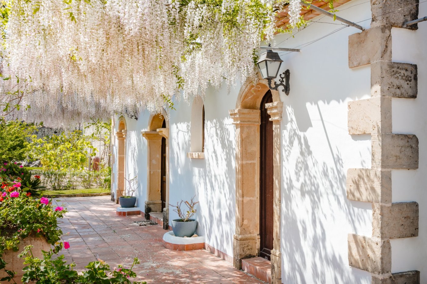 Façade méditerranéenne avec arches en pierre de Tosca et portes en bois, abritée par une pergola de glycines blanches sur un sol en terre cuite.