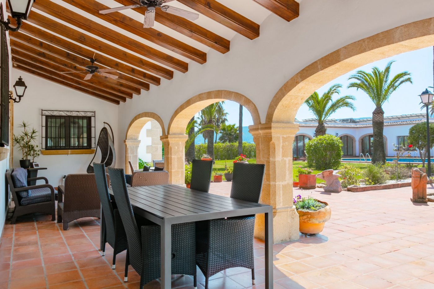 Grande terrasse couverte avec arches en pierre de Tosca, plafonds à poutres apparentes et coin repas offrant une vue sur le jardin méditerranéen et ses palmiers.