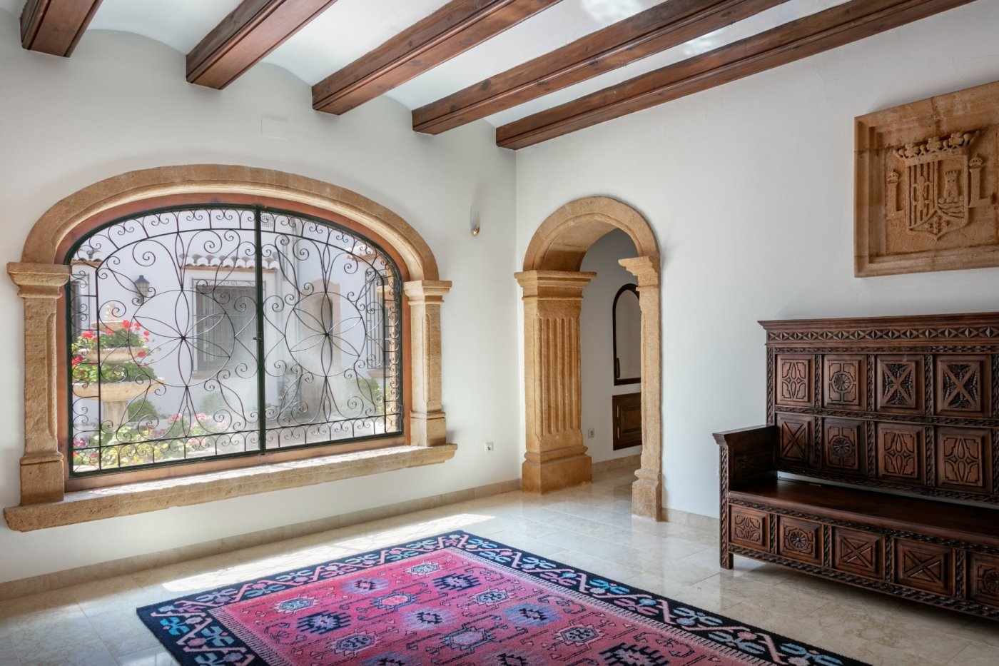 Hall d'entrée avec arches en pierre de Tosca, poutres en bois apparentes et grande fenêtre en fer forgé donnant sur le patio central et sa fontaine.