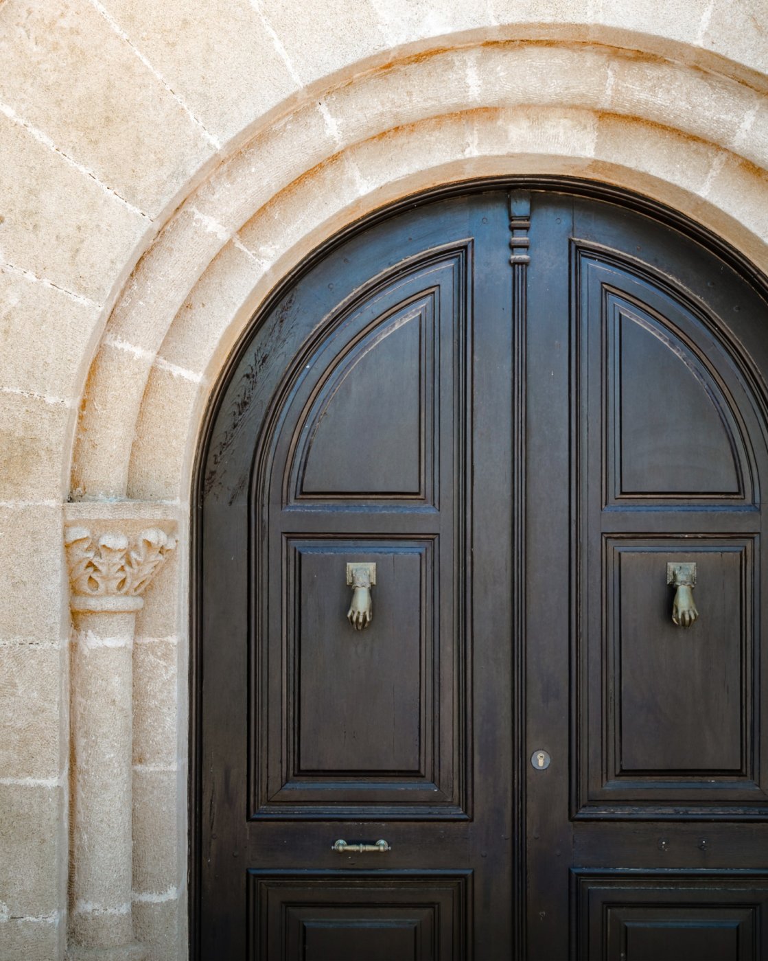 Porte d'entrée cintrée en bois sombre encadrée par une arche en pierre de Tosca, ornée de heurtoirs en laiton et d'une colonne sculptée.