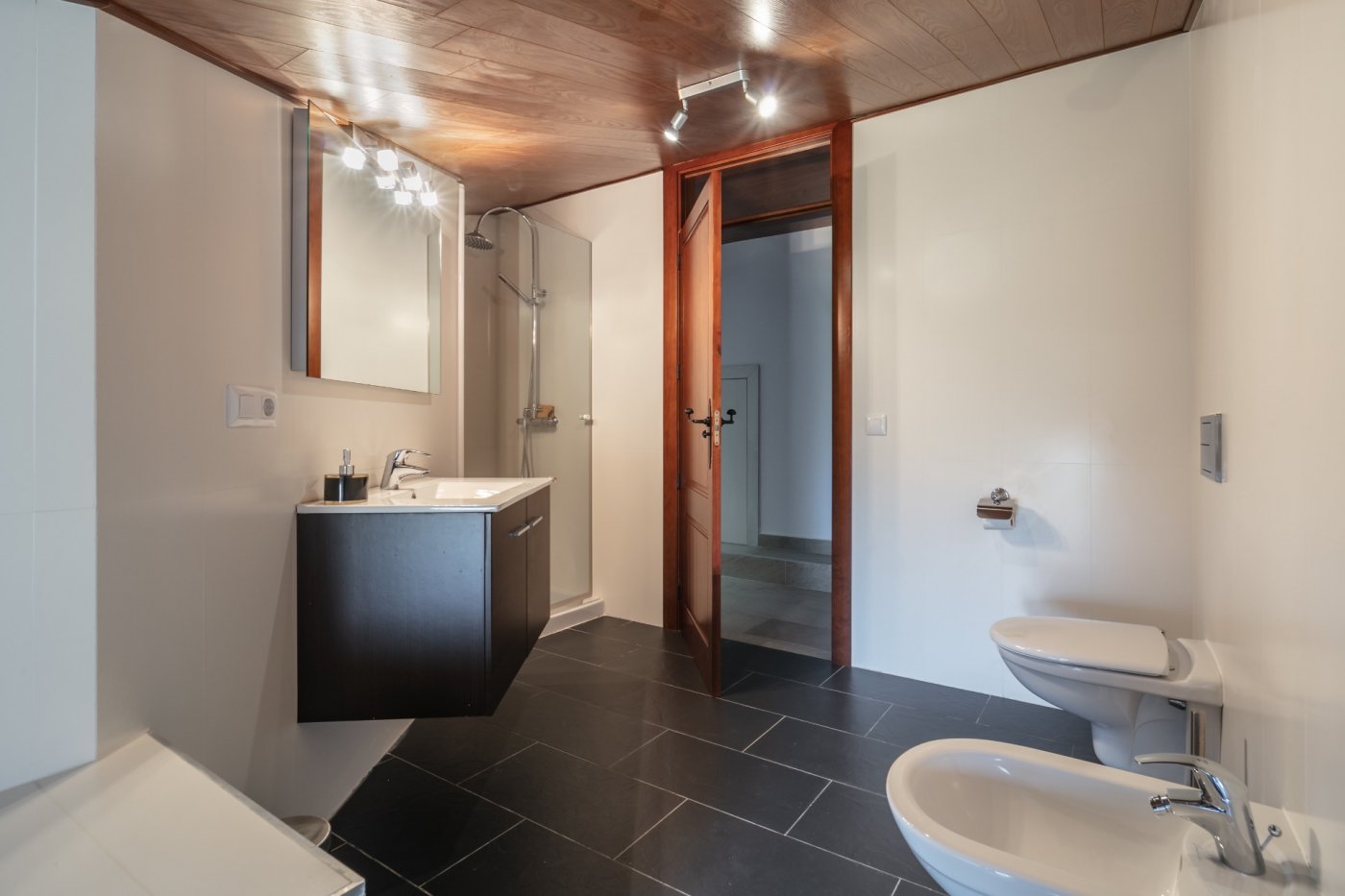 Salle de bain moderne avec plafond en bois, sol en ardoise foncée, meuble vasque suspendu, douche vitrée et sanitaires blancs.