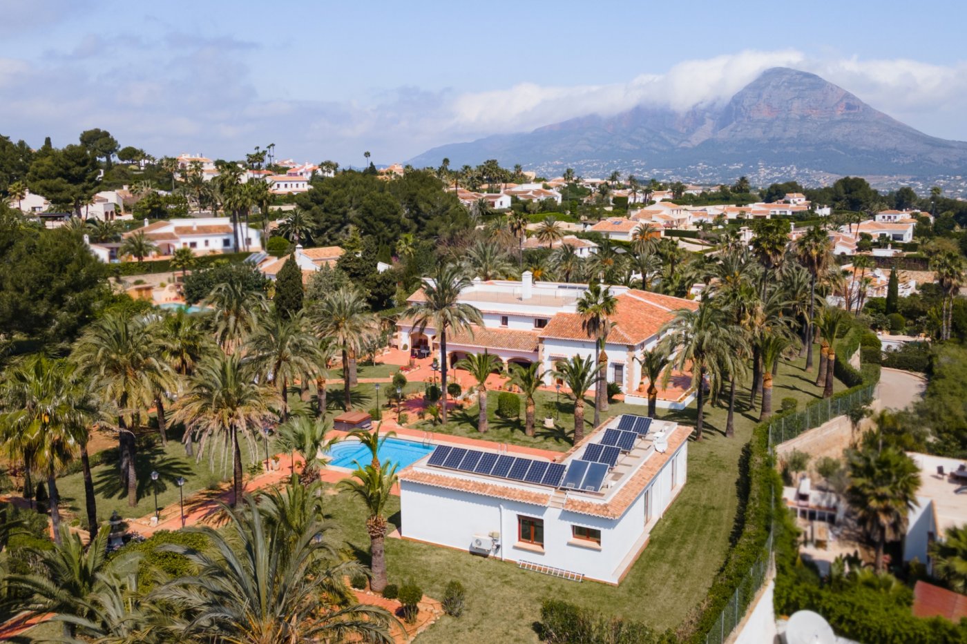 Vue aérienne du domaine comprenant la villa principale, la maison d'amis avec panneaux solaires, la piscine et un vaste jardin de palmiers face au Montgó.