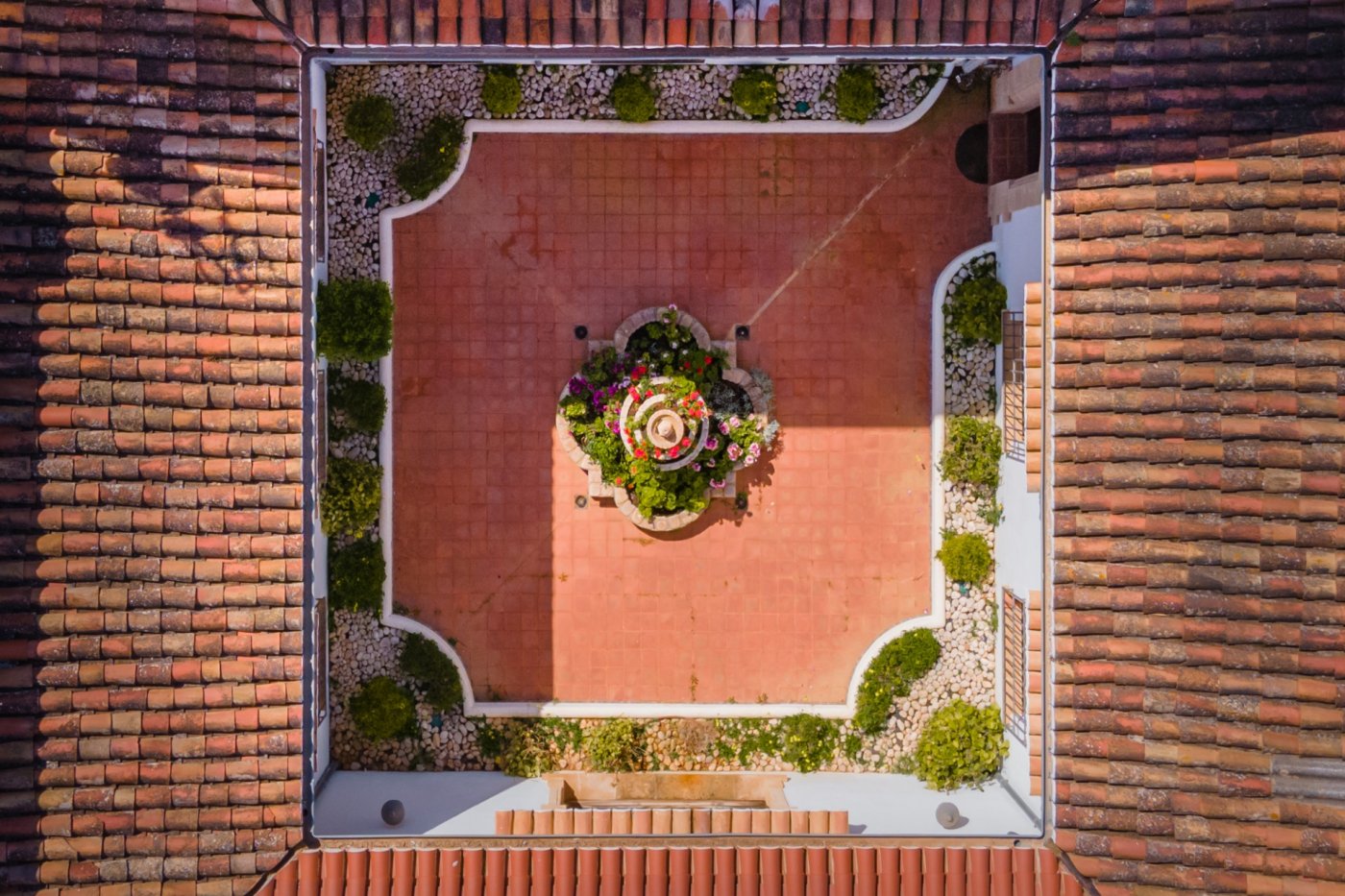Vue aérienne du patio intérieur central avec son sol en terre cuite, sa fontaine fleurie et ses bordures végétalisées sous des toits en tuiles traditionnelles.