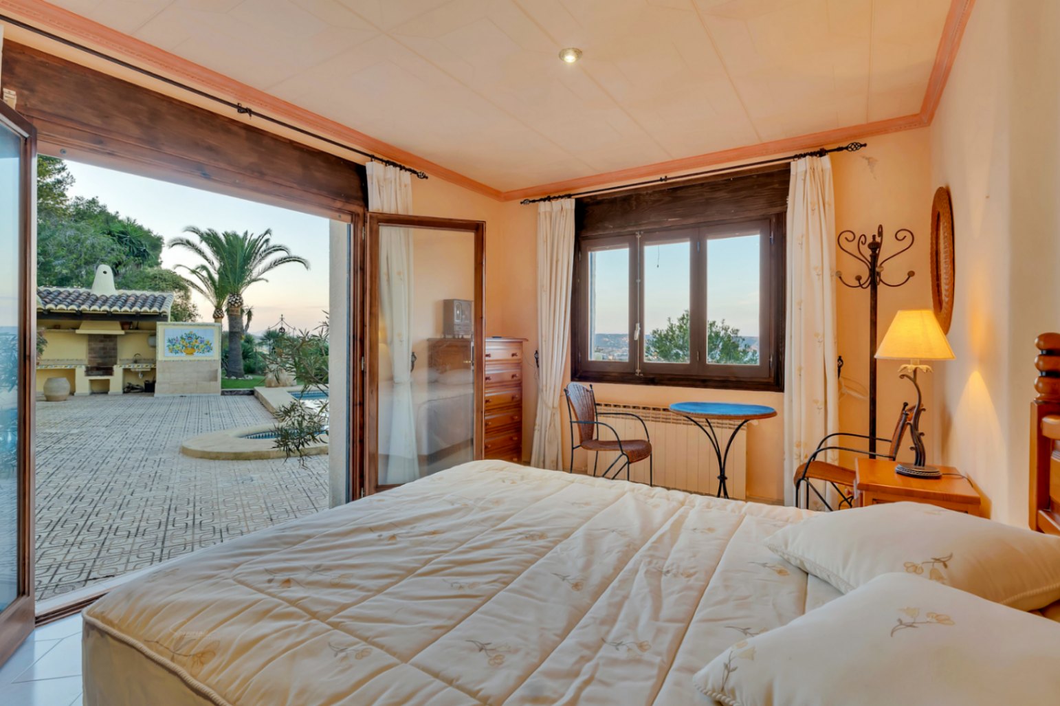 Chambre avec accès direct à la terrasse et la piscine, menuiseries en bois sombre, mobilier classique et vue sur le jardin méditerranéen et l'espace barbecue.
