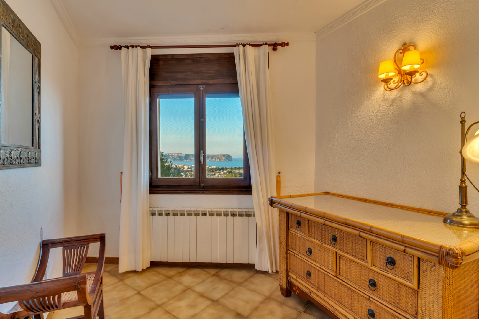 Chambre avec commode en osier, sol en terre cuite et fenêtre en bois offrant une vue sur la mer. Équipée d'un radiateur et d'une applique murale élégante.