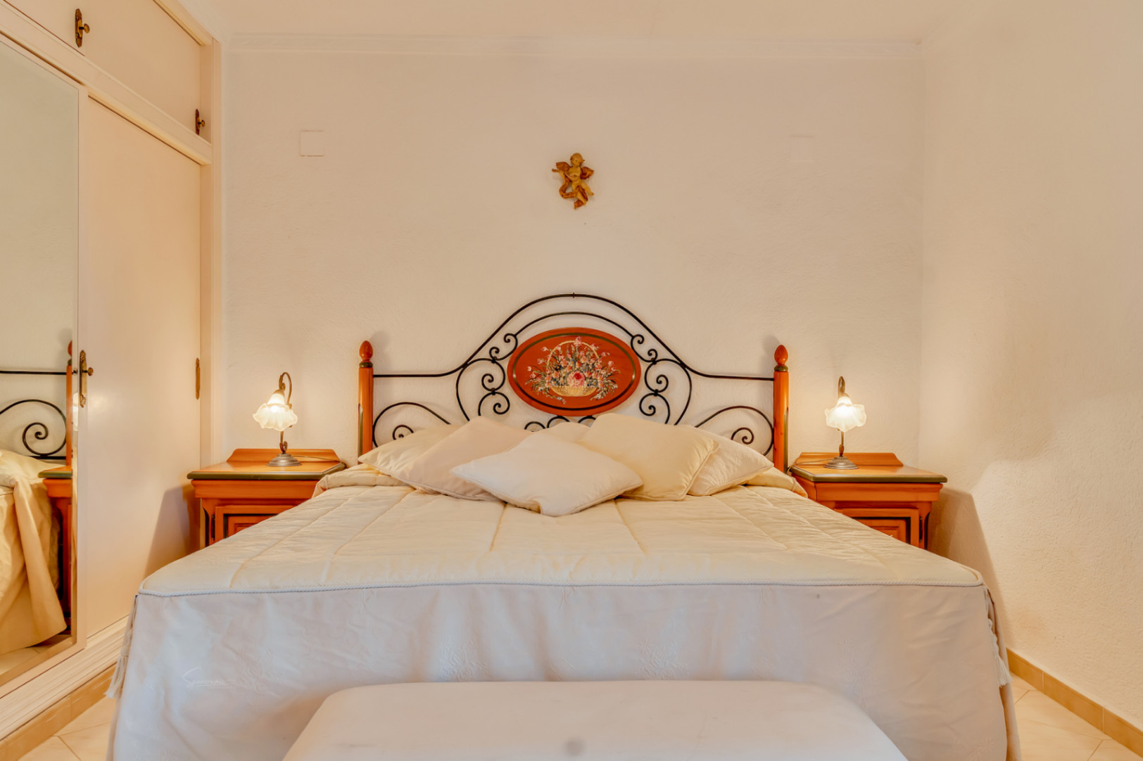 Chambre avec lit double, tête de lit en fer forgé à motif floral, tables de chevet en bois et placards intégrés avec porte miroir.