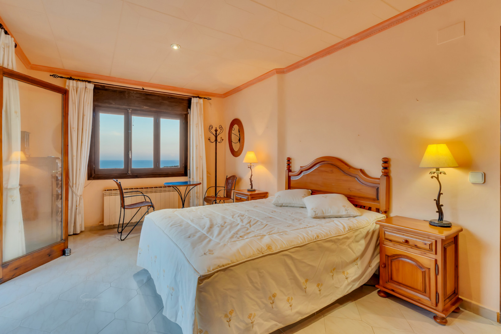 Chambre avec mobilier en bois classique, sol en carrelage hexagonal et fenêtre offrant une vue sur la mer. Équipée d'un radiateur et d'un petit coin salon.