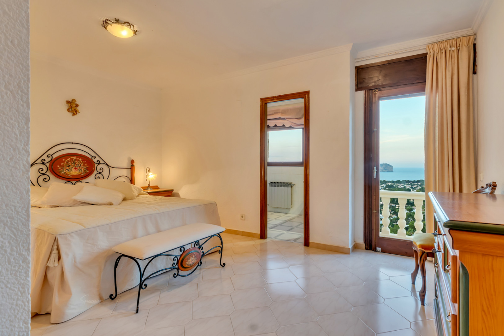 Chambre avec tête de lit en fer forgé, carrelage clair et porte vitrée ouvrant sur une terrasse avec vue sur la mer et le Cabo San Antonio.