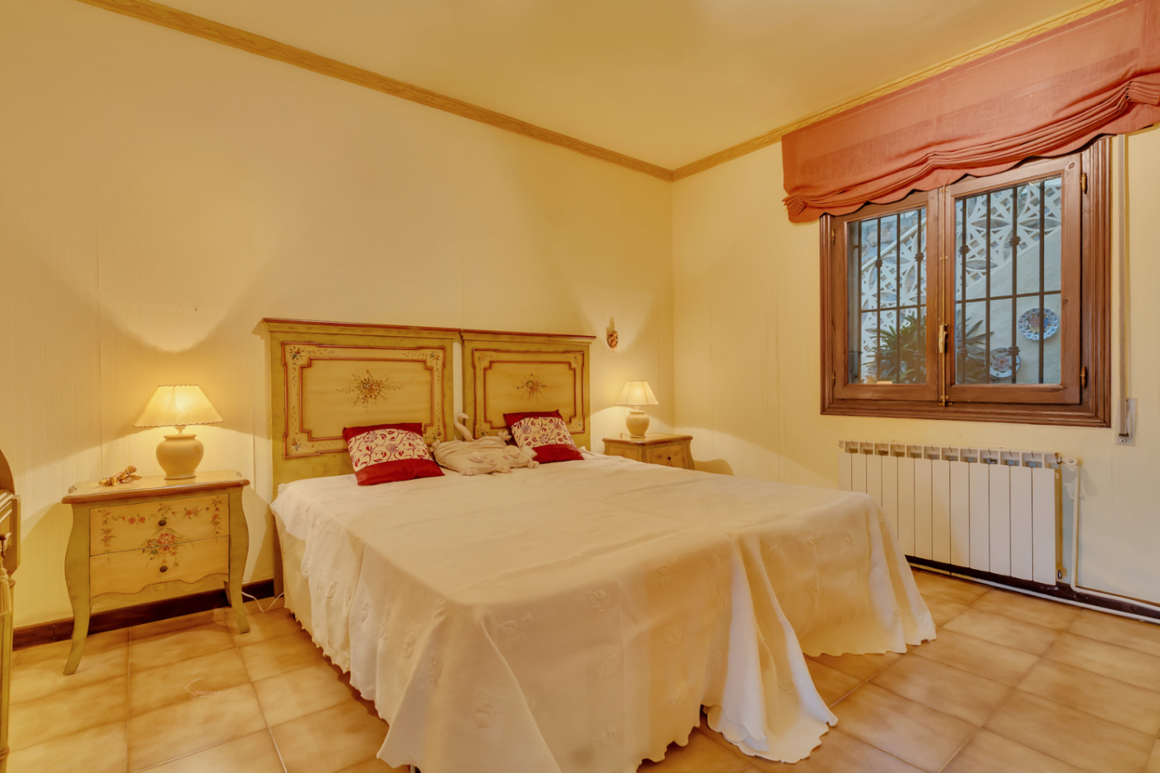 Chambre double avec têtes de lit en bois peintes à la main, tables de chevet assorties, sol en terre cuite et fenêtre avec cadre en bois et radiateur.