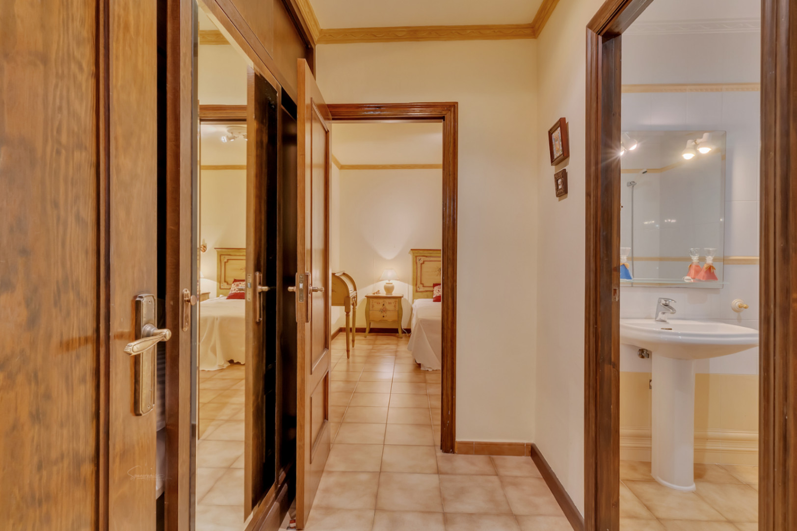 Couloir avec sol en grès reliant une chambre à lits jumeaux, une salle de bain avec lavabo sur colonne et des placards intégrés en bois munis de miroirs.