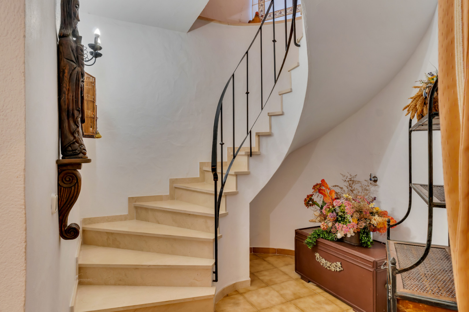 Escalier courbe avec marches en pierre claire et rampe en fer forgé noir. L'espace comprend des murs blancs et un sol en carrelage céramique traditionnel.