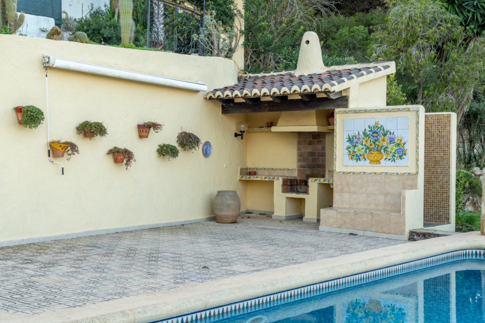 Espace barbecue maçonné avec toit en tuiles, fresque décorative en céramique et douche extérieure à côté de la piscine sur terrasse pavée.