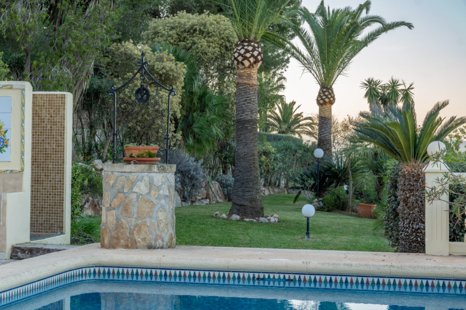 Jardin méditerranéen avec pelouse, palmiers et puits décoratif en pierre près de la piscine. Comprend une douche extérieure carrelée et des luminaires sphériques.