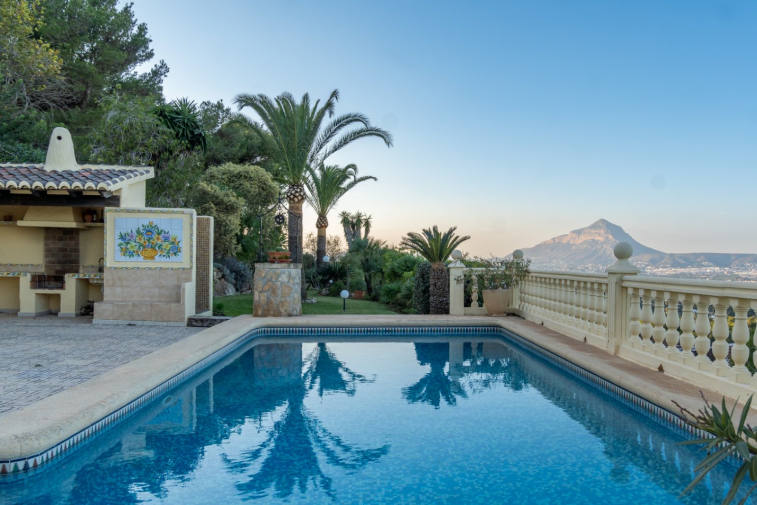 Piscine en mosaïque bleue avec cuisine d'été couverte et décor en céramique, offrant une vue dégagée sur le Montgó et la végétation locale.