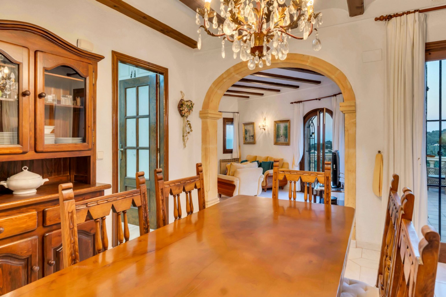 Salle à manger avec mobilier en bois massif, poutres apparentes et arche en pierre de Tosca ouvrant sur le salon et la terrasse.