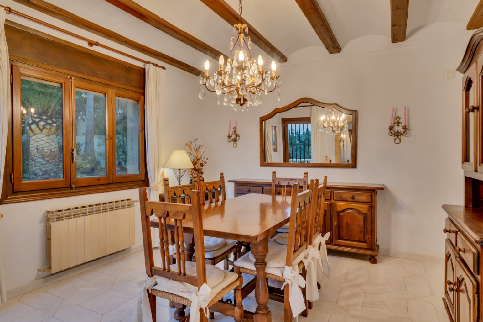 Salle à manger traditionnelle avec poutres en bois, mobilier en bois massif, lustre en cristal et sol en carrelage clair aux motifs géométriques.