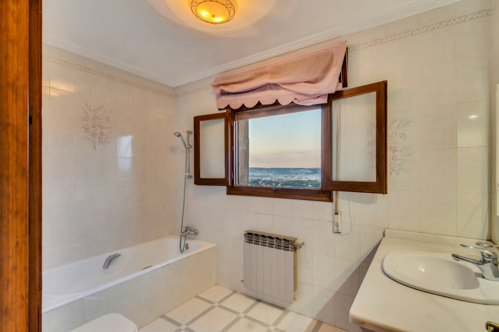 Salle de bain avec baignoire, carrelage à motifs floraux et fenêtre en bois offrant une vue panoramique sur la mer et la baie.
