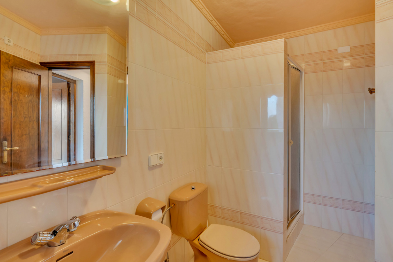 Salle de bain avec carrelage crème, sanitaires de couleur pêche et douche avec paroi vitrée. Finitions classiques et grand miroir reflétant la porte en bois.