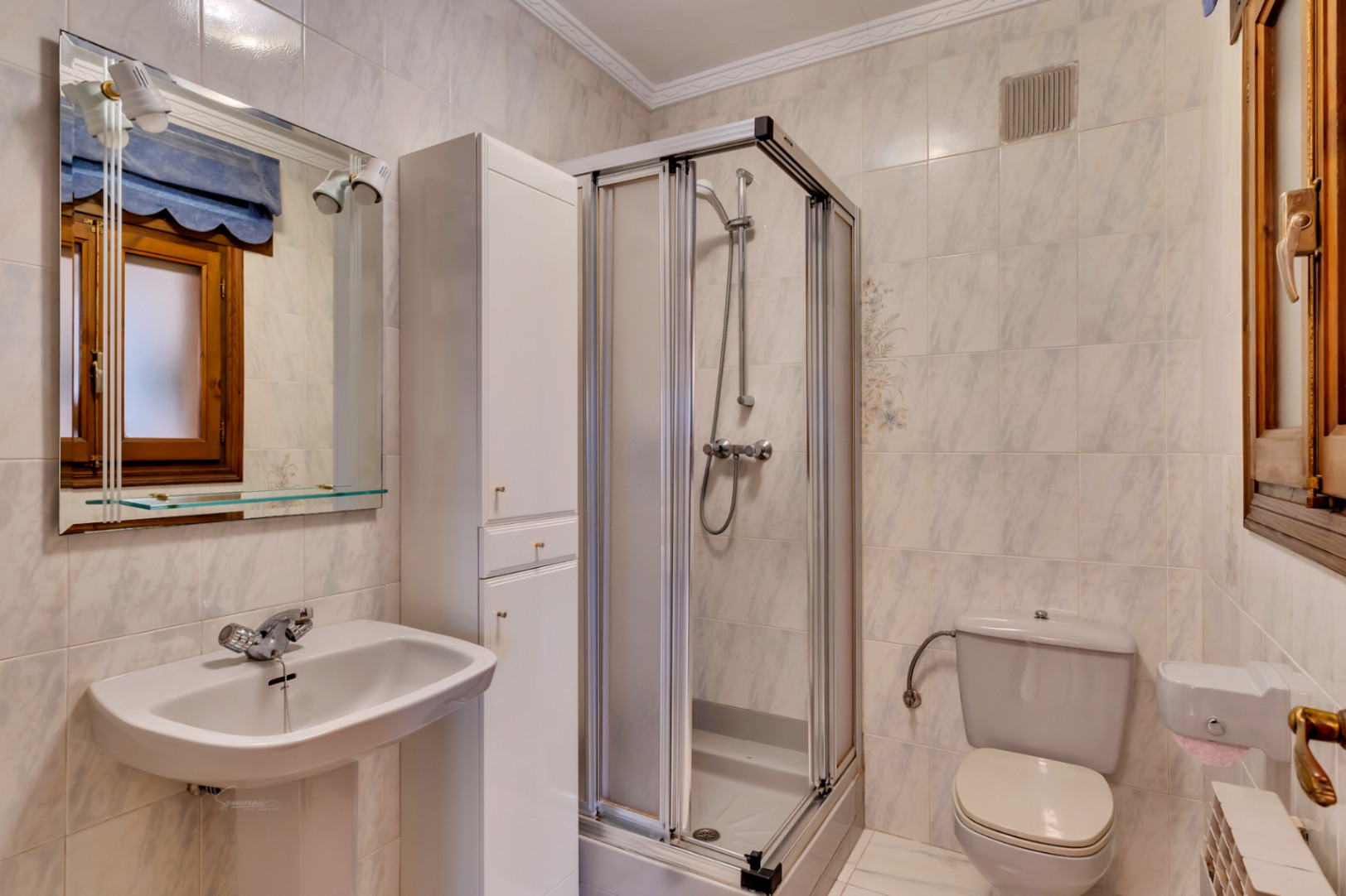 Salle de bain équipée d'un lavabo sur colonne, d'une armoire de rangement blanche et d'une cabine de douche avec parois coulissantes.