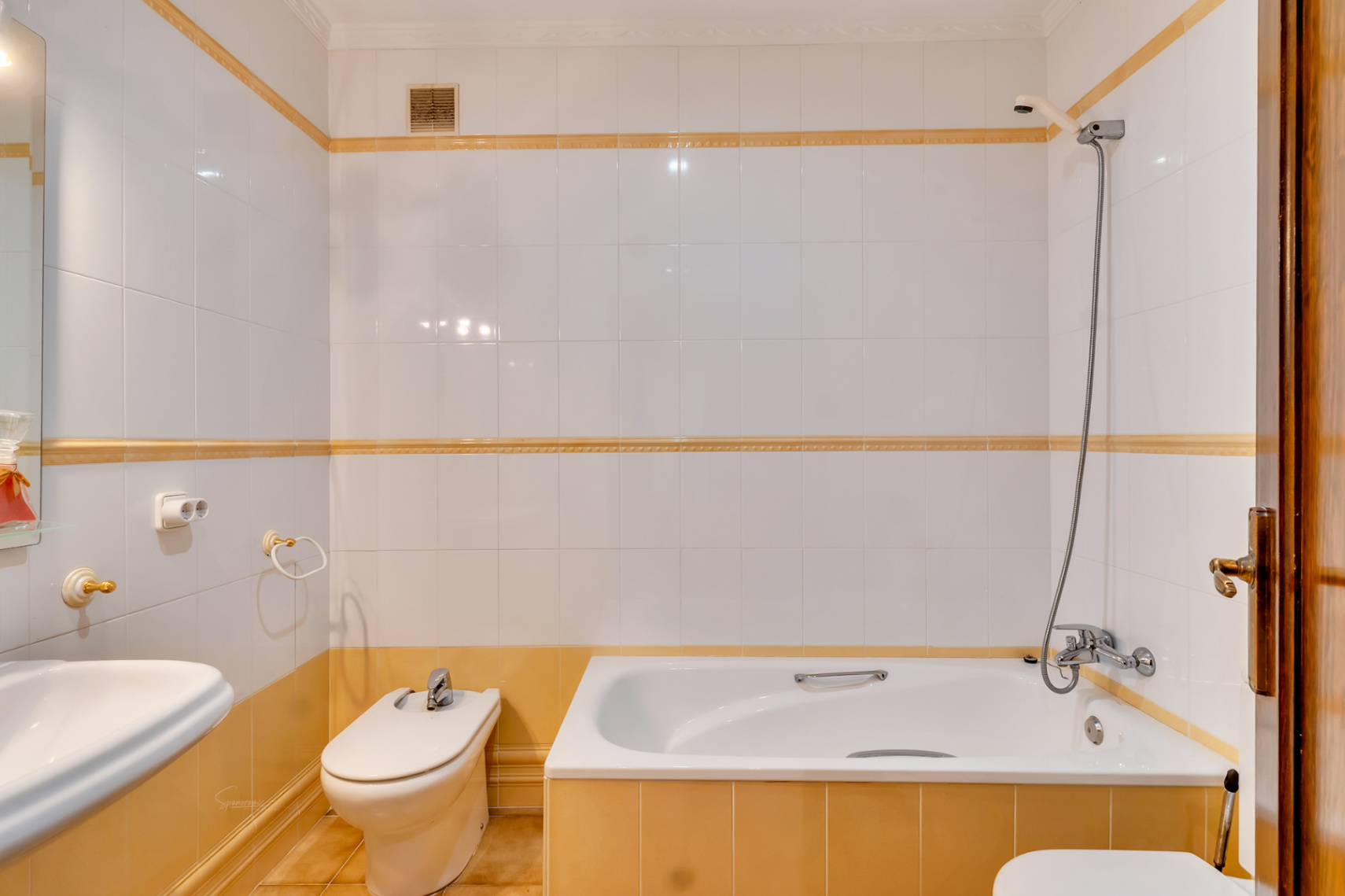 Salle de bains avec carrelage blanc et liserés ocres, comprenant une baignoire, un bidet et une robinetterie classique. Les finitions incluent des accessoires dorés.