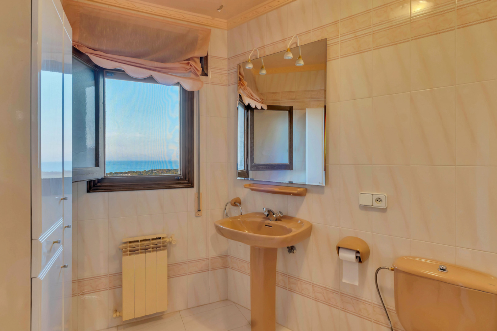Salle de bains avec sanitaires de couleur crème, lavabo sur colonne et fenêtre offrant une vue dégagée sur la mer et le littoral.