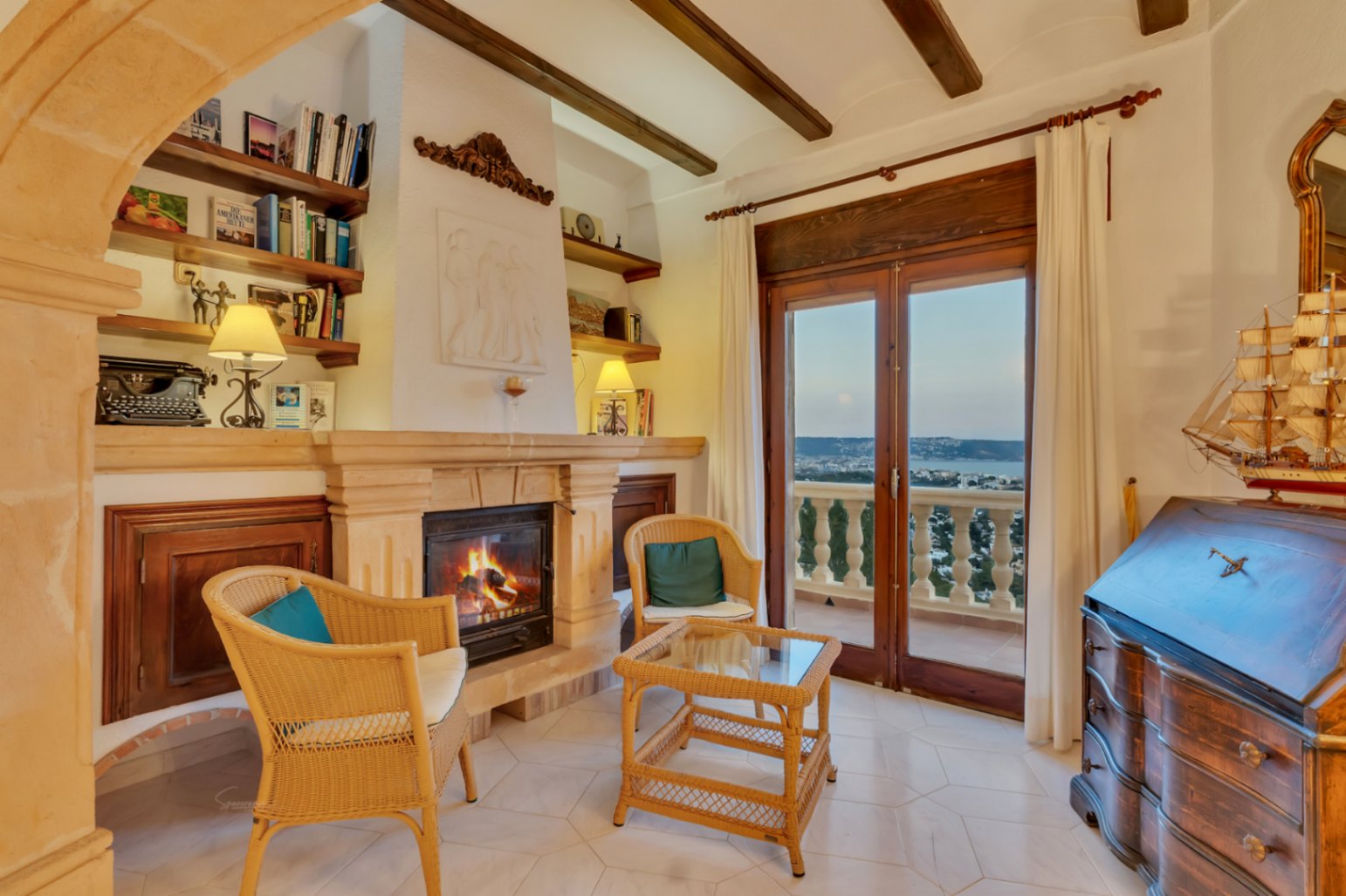 Salon avec cheminée en pierre, poutres apparentes et mobilier en osier. Les portes vitrées offrent une vue sur la mer et un accès direct à la terrasse.