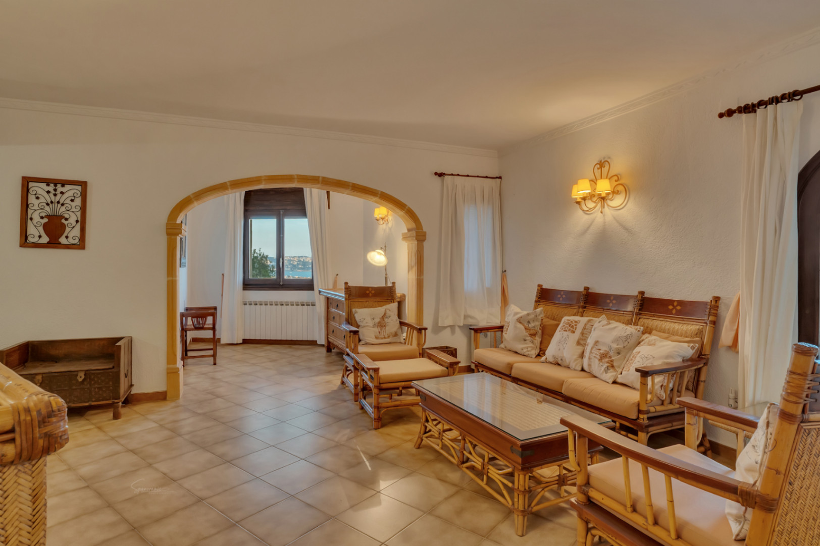 Salon avec mobilier en bambou et arche en pierre de Tosca. L'espace dispose de sols en céramique et d'une ouverture sur une pièce avec vue sur la baie de Jávea.