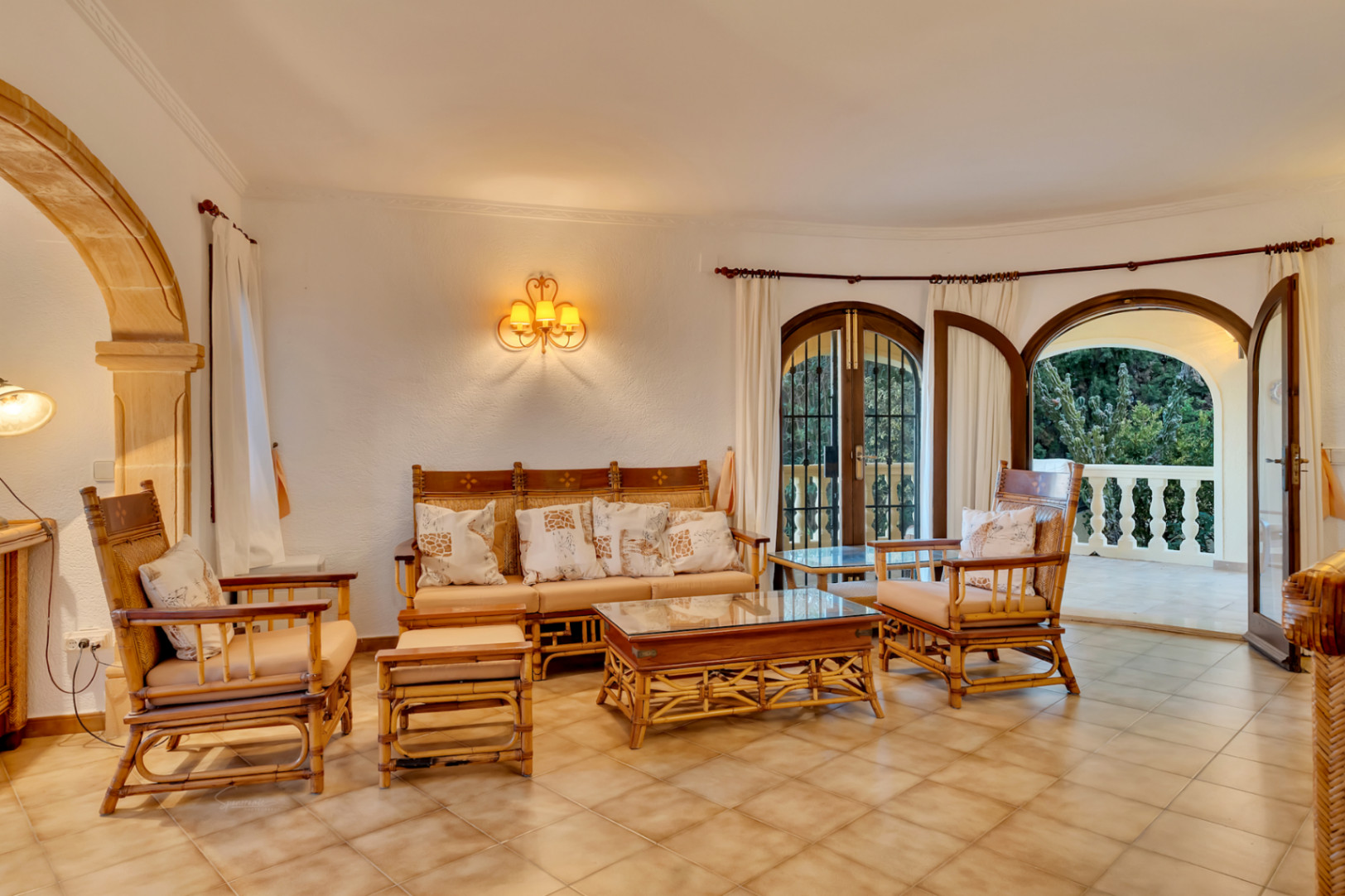 Salon avec mobilier en bambou, sol en céramique et arche en pierre de Tosca. Des portes cintrées en bois mènent à une terrasse avec balustrade blanche.