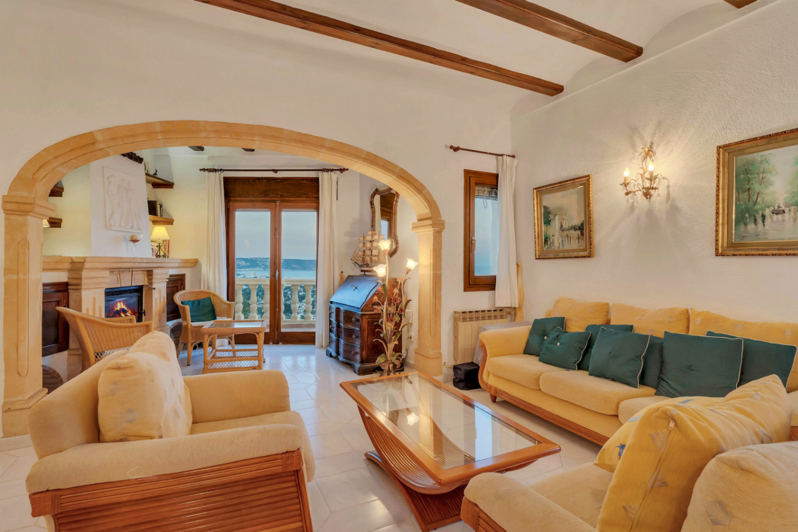 Salon de style méditerranéen avec poutres en bois apparentes, arche en pierre de Tosca et cheminée. Accès direct à une terrasse offrant une vue sur la mer.