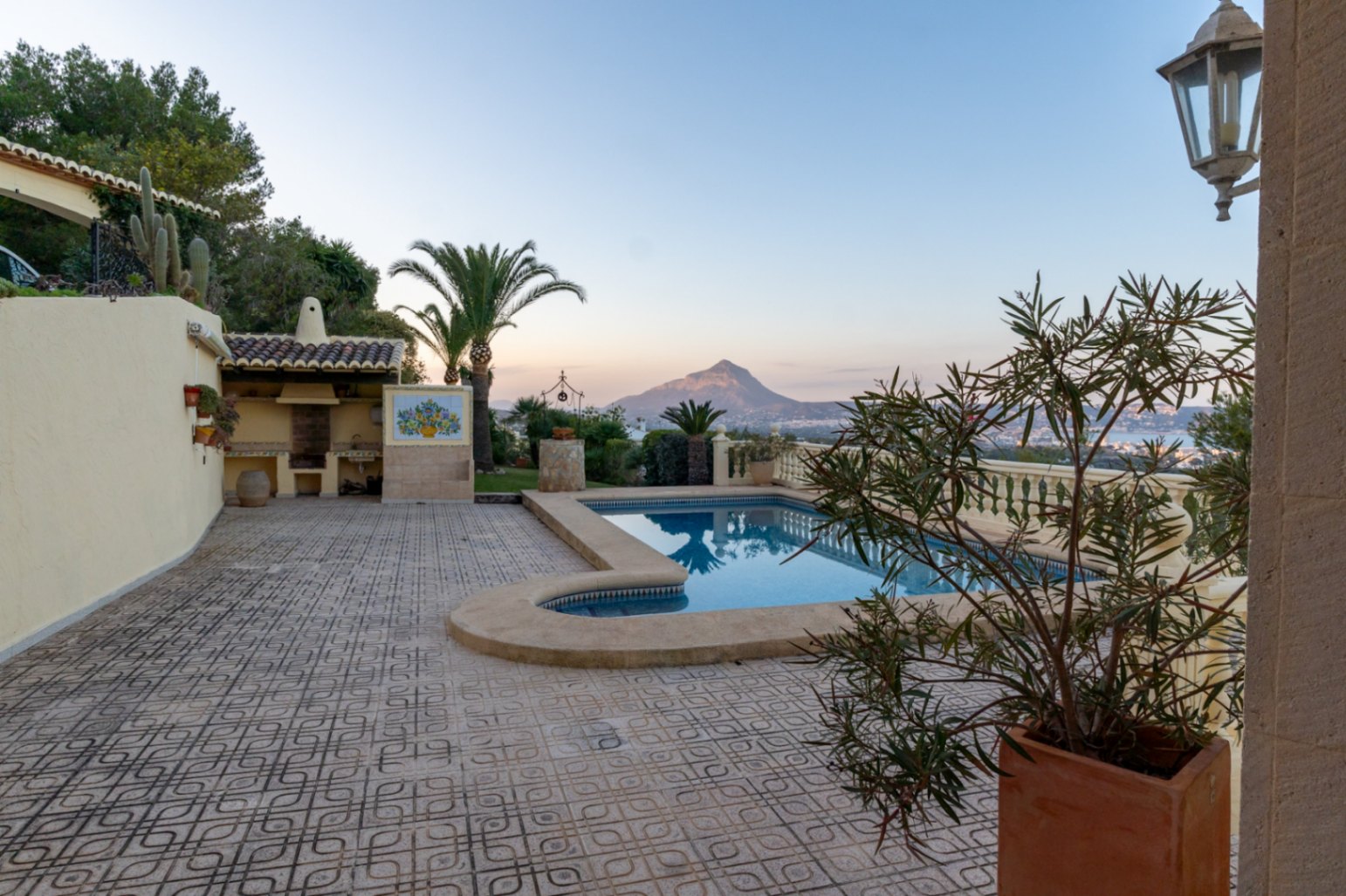 Terrasse extérieure avec carrelage ornemental, piscine rectangulaire et espace barbecue couvert. Vue dégagée sur le Montgó et la baie à la tombée du jour.