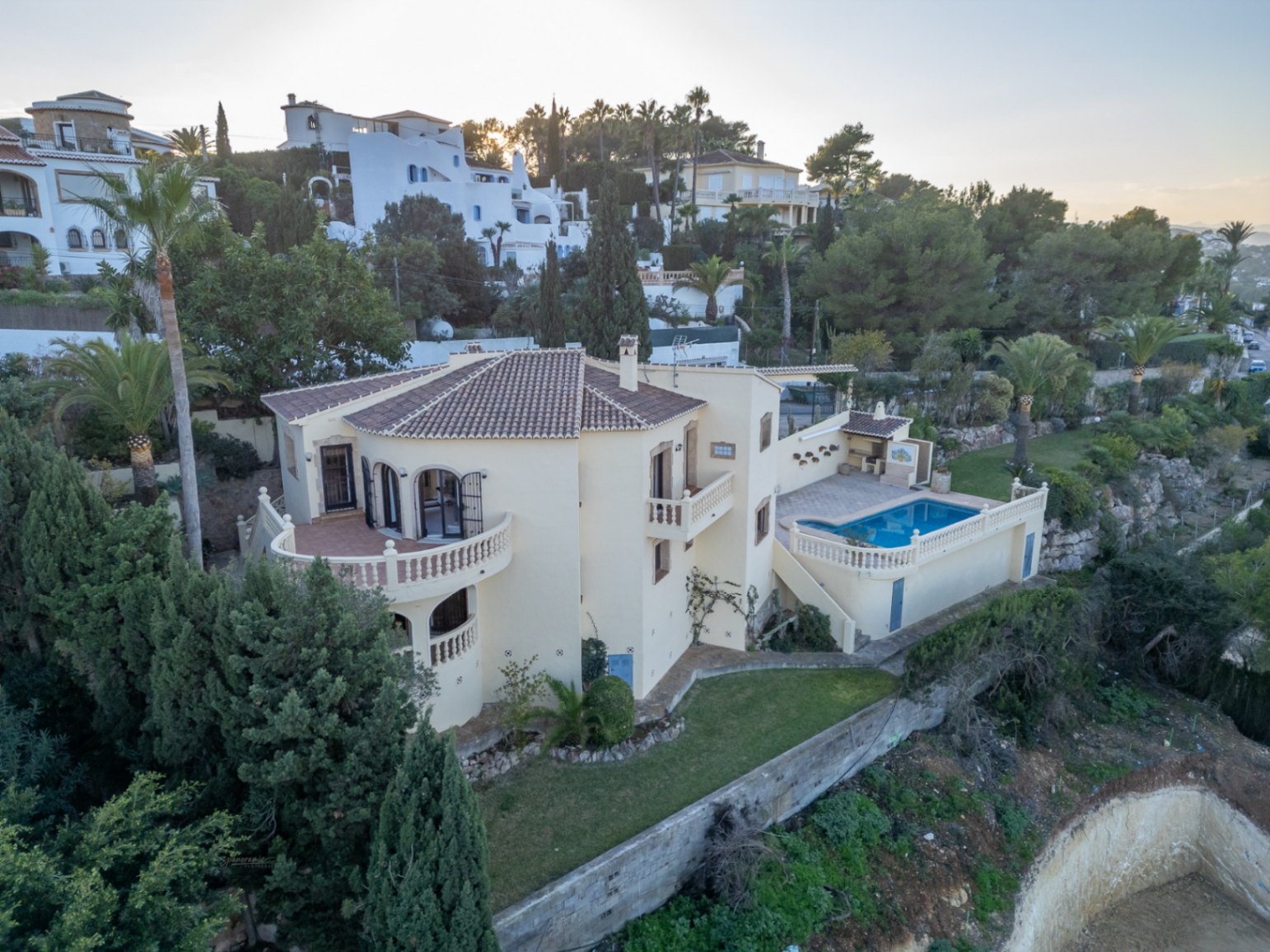 Vue aérienne de la villa avec façade crème, toit en terre cuite et terrasses à balustrades. L'extérieur dispose d'une piscine, d'un barbecue et d'un jardin arboré.