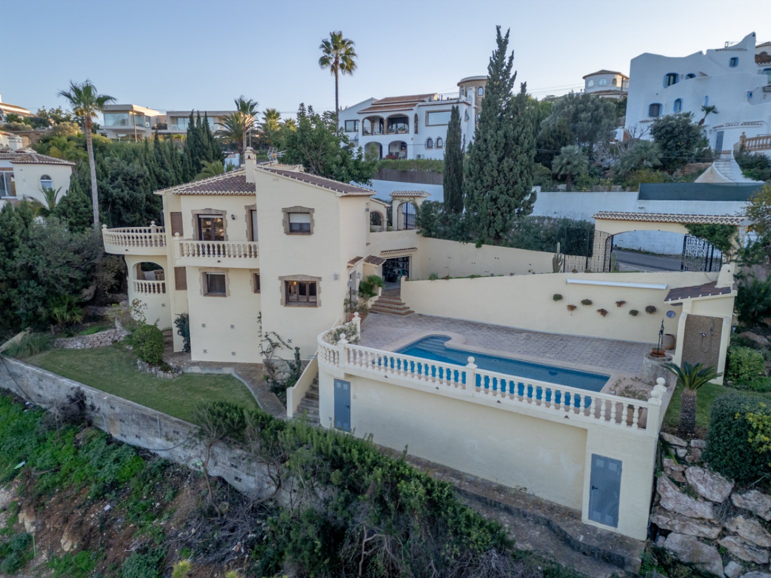 Vue extérieure de la villa méditerranéenne avec piscine privée, terrasses à balustrades blanches et façade crème ornée de cadres de fenêtres en pierre.