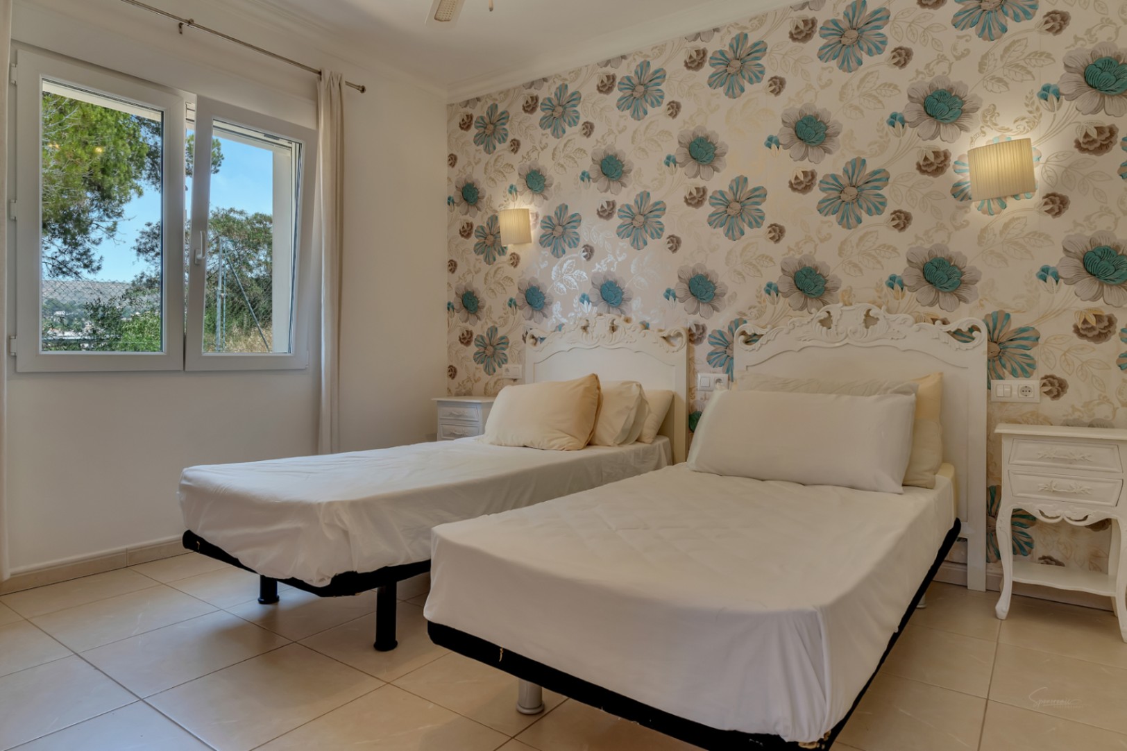 Chambre avec deux lits simples, têtes de lit blanches ornementées et papier peint floral. Comprend un sol en carrelage et une fenêtre ouvrant sur la nature environnante.