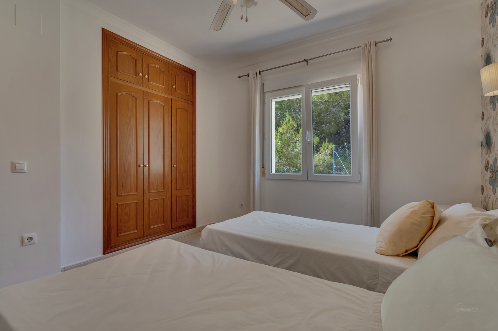 Chambre avec deux lits simples, une grande armoire encastrée en bois et une fenêtre donnant sur les pins. Comprend un ventilateur de plafond et un mur tapissé.