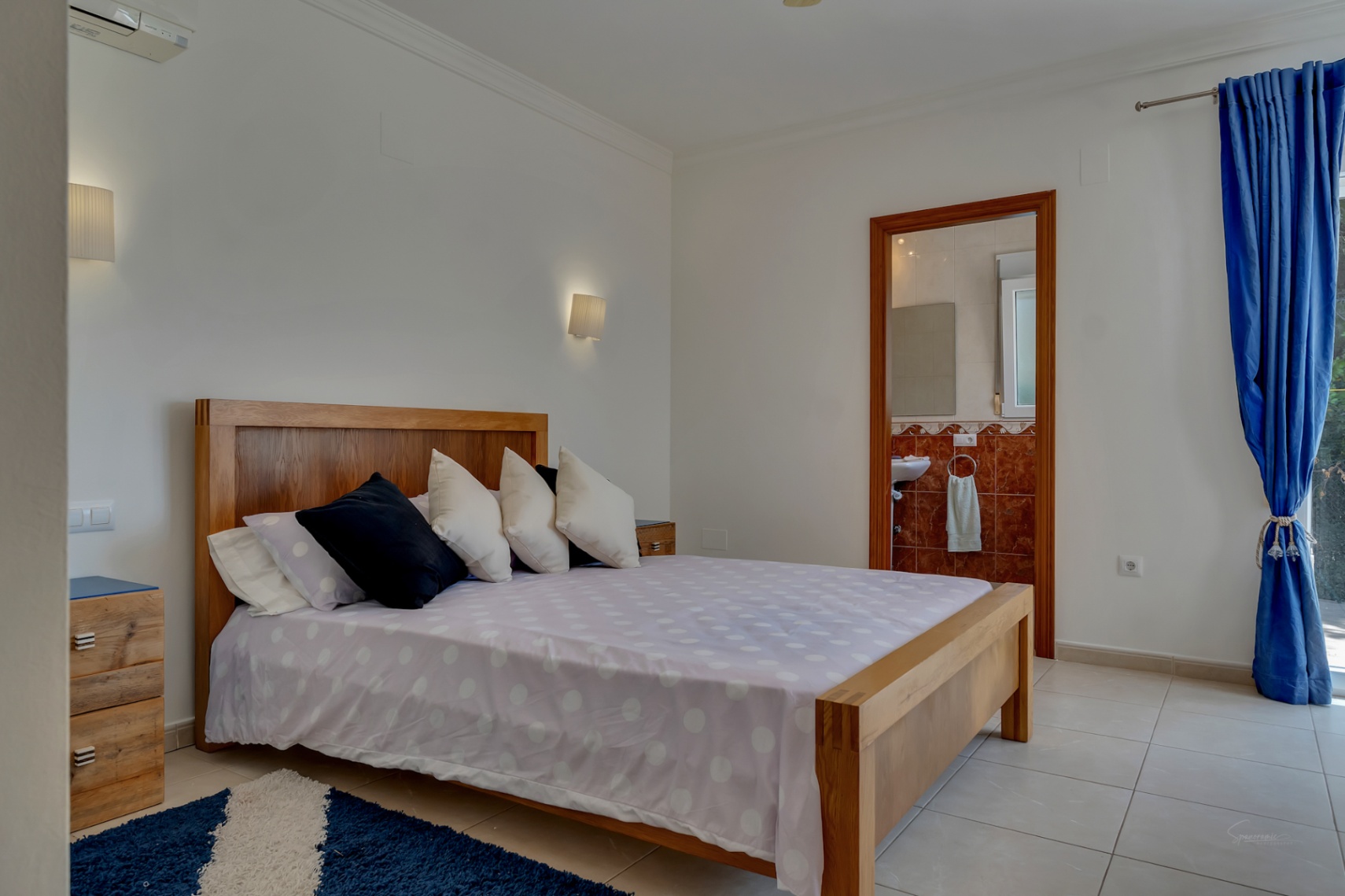 Chambre lumineuse avec cadre de lit en bois massif, sol en grès, climatisation et salle de bain attenante avec revêtement en marbre rouge.