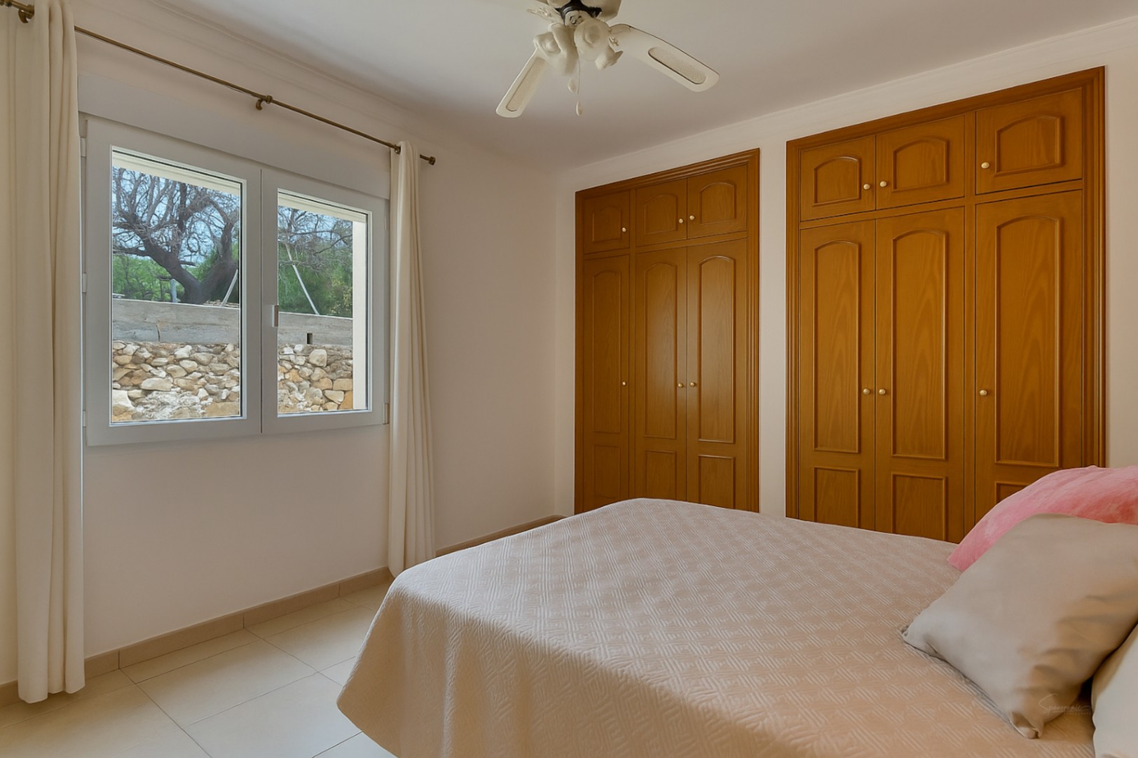 Chambre lumineuse avec placards intégrés en bois, ventilateur de plafond et fenêtre offrant une vue sur le jardin et un mur en pierre.