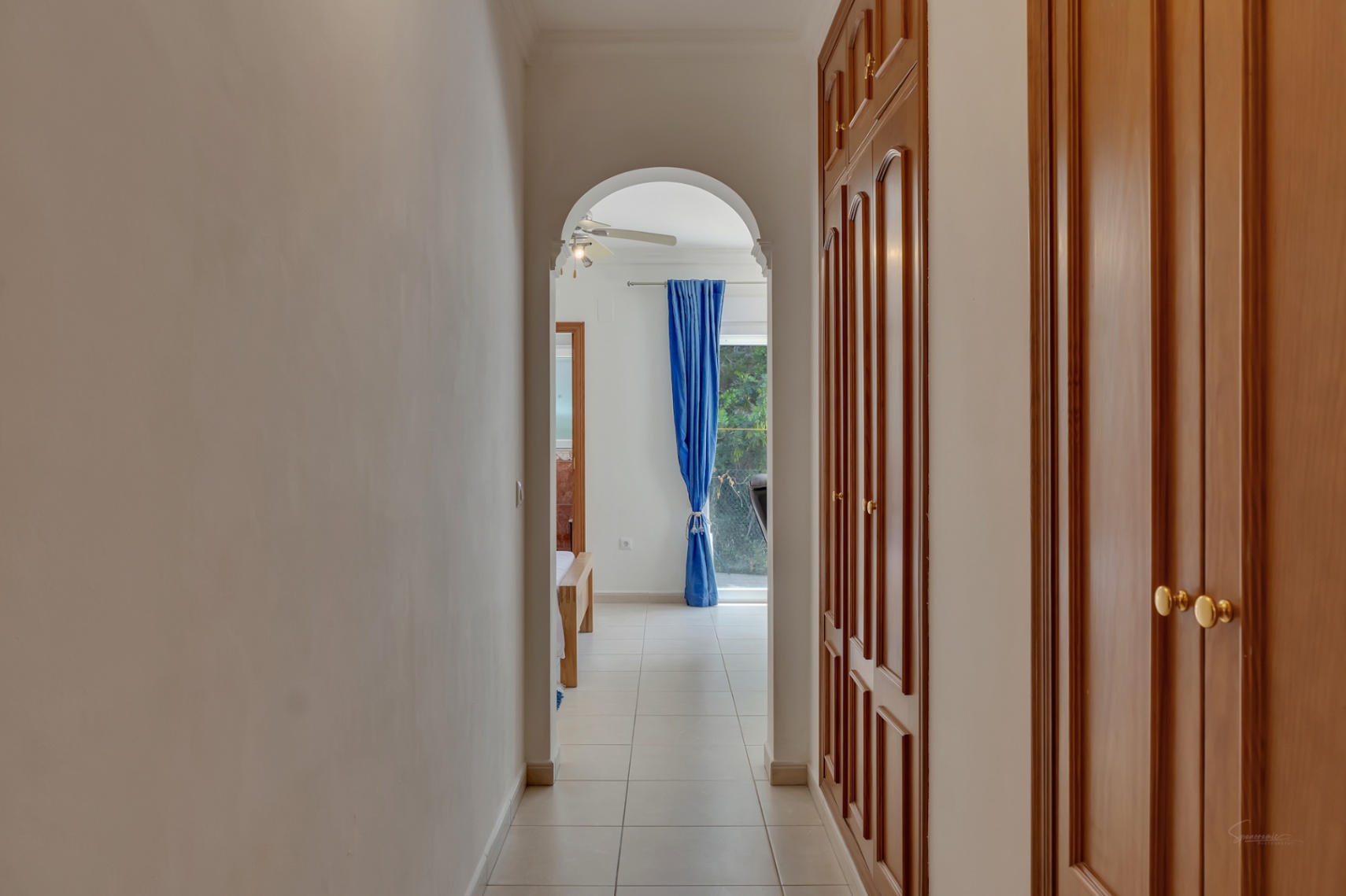 Couloir doté de grands placards intégrés en bois et d'un sol en céramique, menant par une arche à une chambre lumineuse avec ventilateur de plafond.